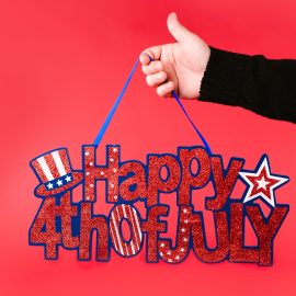 4th of July Bike Parade & Community Event A hand holding a glittery 'Happy 4th of July' sign against a vibrant red background.